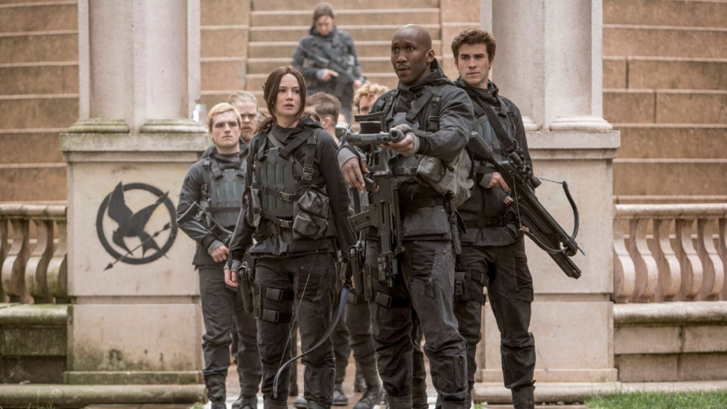 A still from The Hunger Games film. A group of actors stand wearing Throup's clothing. They're holding guns and dressed all in black.