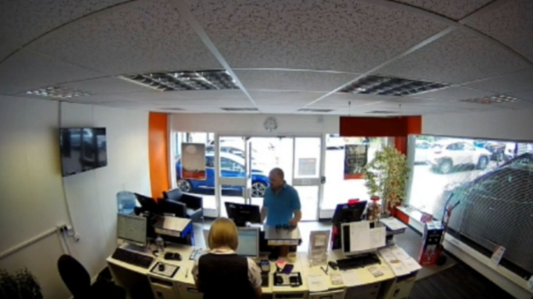 CCTV of Stewart Ahearne dressed in a blue polo shirt at the counter of an Avis hire company as he attempts to hire out a vehicle.