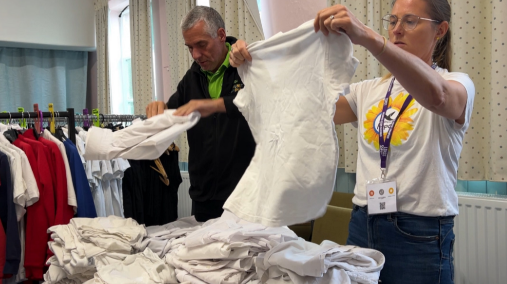 Volunteers sorting through the uniforms. A man with short grey hair and wearing a green t-shirt underneath a black jumper is folding a white shirt. Next to him is a woman with light brown hair in a pony tail and with glasses. She is holding up a white shirt. In front of them is a pile of white shirts and to the side is a clothes rail with various items on.