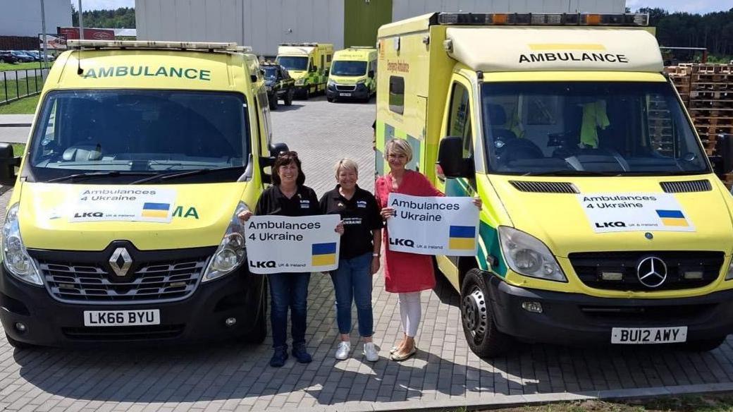 Three women are standing between two yellow ambulances that are park in front of large white buildings. Two of the women are holding up posters that state: "Ambulances 4 Ukraine." 