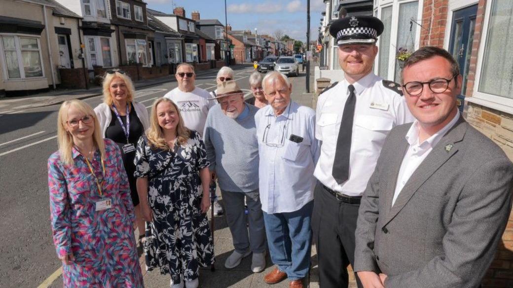 In the right is Sunderland City Council Leader Cllr Michael Mordey with Superintendent Mark Hall. From left: Deputy Leader of Sunderland City Council and Cabinet Member for Health, Wellbeing and Community Safety, Councillor Kelly Chequer, and second from left in the foreground Cabinet Member for the Environment, Councillor Lindsey Leonard with Hendon residents. They are smiling and standing on the pavement. The street behind them has rows of terraced houses and a number of parked cars.