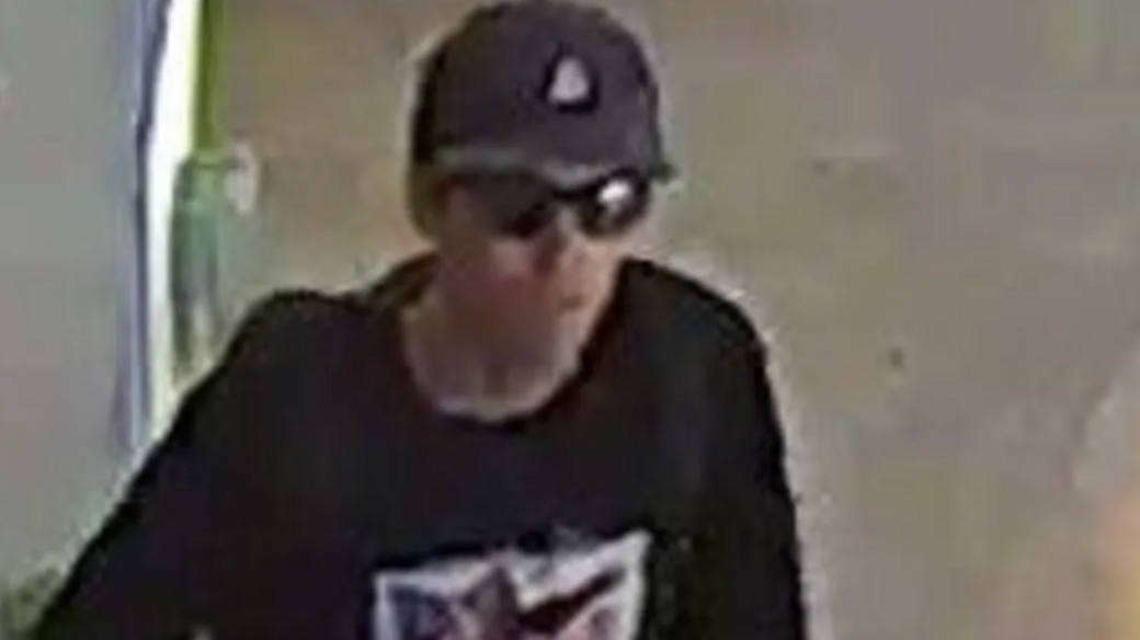 Grainy photo taken from video of young white man with gray-black hat with triangle logo, black sunglasses, black T-shirt