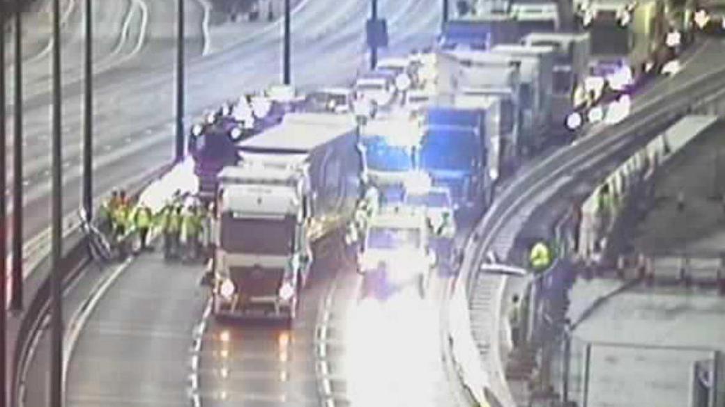 A traffic camera showing emergency vehicles on the M25. A lorry is at the front and in the background are vehicles queuing.