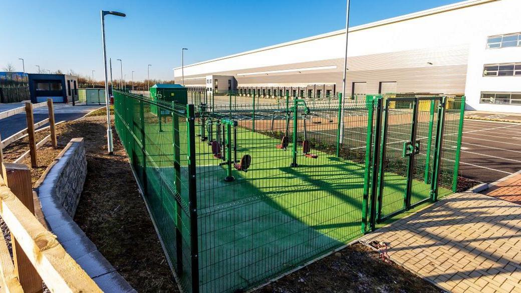 Gym area surrounded by green metal grid fence. Exercise equipment is visible within including seesaw-like device