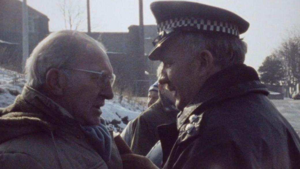 Man with grey hair and glasses to the left of frame is wearing a large coat and is facing off with a police officer. The officer is wearing his uniform which includes his black and white flat cap and a black overcoat. Around them are a few other miners who are standing on the picket line.