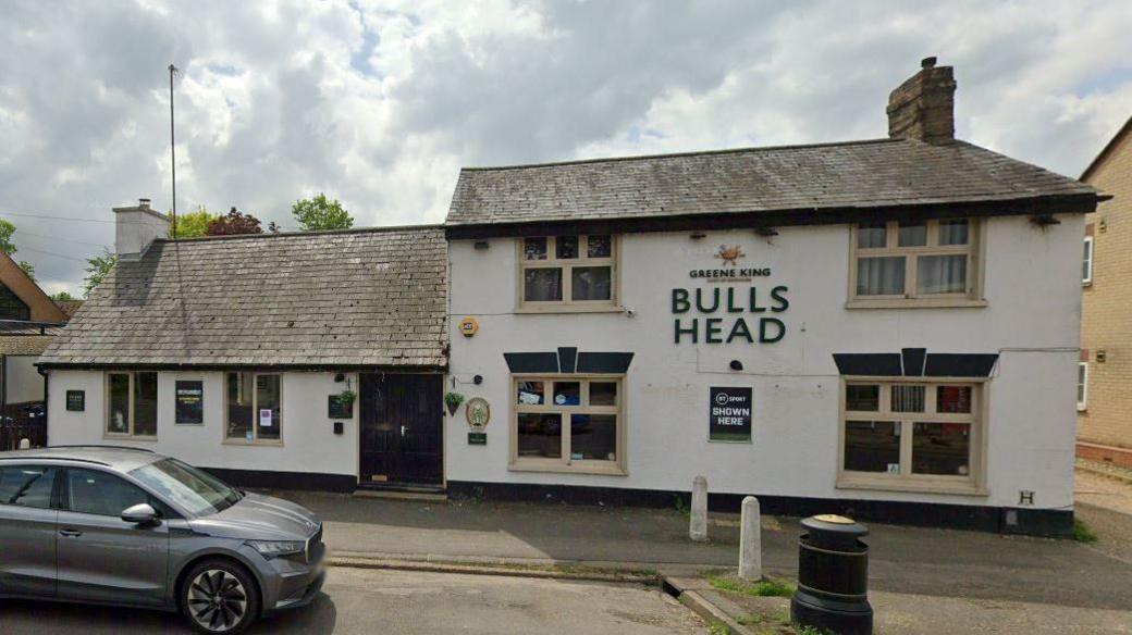 The exterior of the Bulls Head pub in St Neots, likely before it closed down. It is a white two-storey building on the right, with a single storey building on the left and a black door for its entrance. Bulls Head is written in large letters in the middle of the two-storey building. In front of it is a pavement and a grey car.