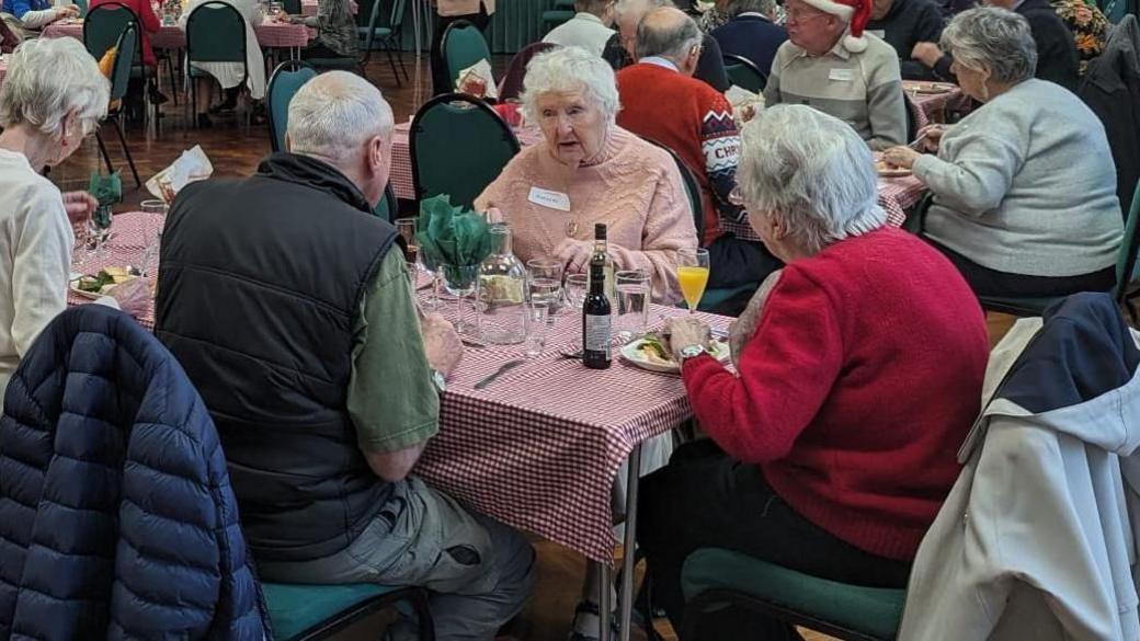 A group of elderly people sat around a table with glasses of wine and plates of food