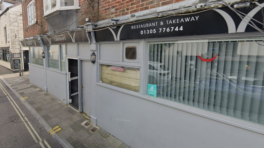 A general view of the Weymouth Tandoori restaurant, which has a grey wall and windows around its outer edge. It is on a town street, with a small pavement in front of it 