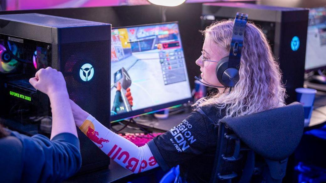 A woman with blonde hair looks as screen while wearing a gaming headset. She fist bumps a player next to her while a game plays on the screen in front of her