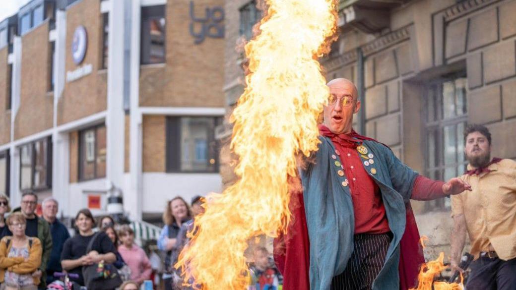 A fire artist is performing to a crown standing behind him. He is a bald man with a red shirt and blue jacket on and there is a large ball of flame in front of him
