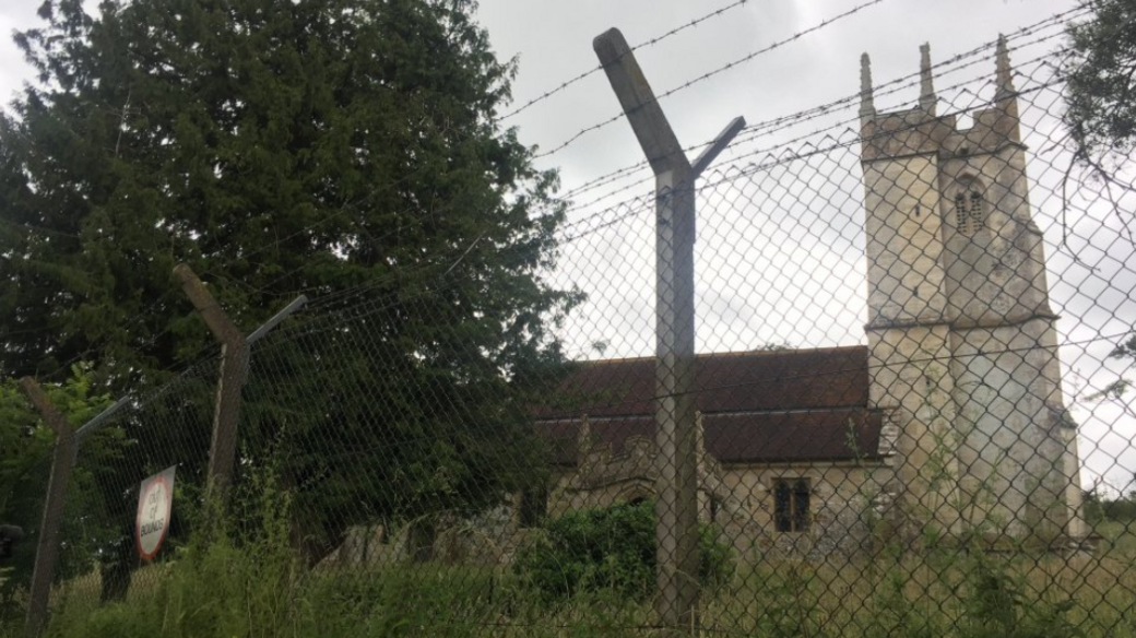 A church with a tower stands behind a tall mesh wire fence with rows of barbed wire at the top