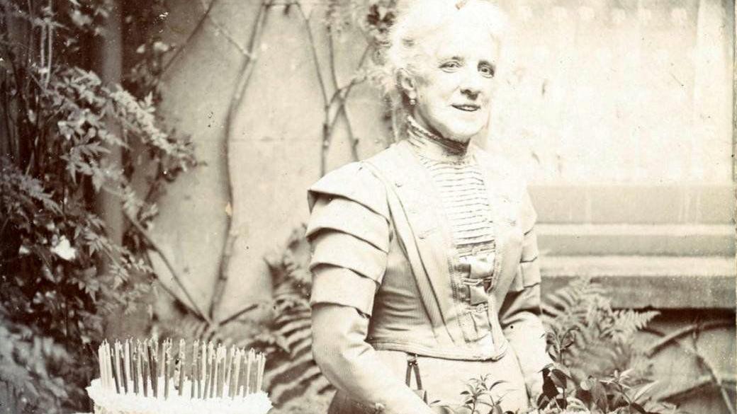 A black and white image of an elderly woman in an old-fashioned bodiced dress standing next to a birthday cake that has dozens of long candles.