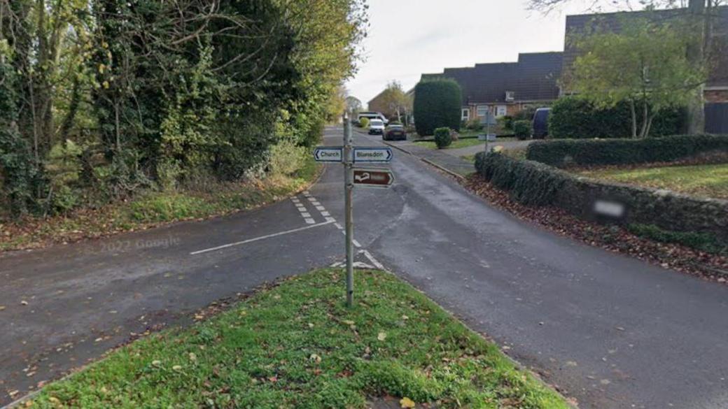 A picture taken on Blundson High Street shows street signs pointing to the right for the church and to the left for Blundson. In the background, there is a nice and quiet residential street with only a few cars parked on the pavement, and plenty of green trees and bushes.