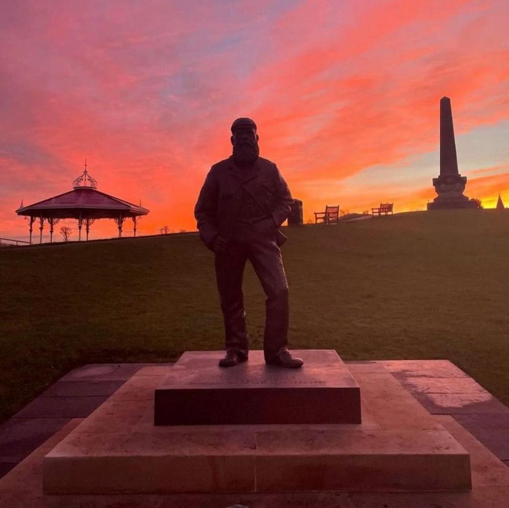 The statue along with a bandstand and monument are silhouetted against a pink and orange sky.