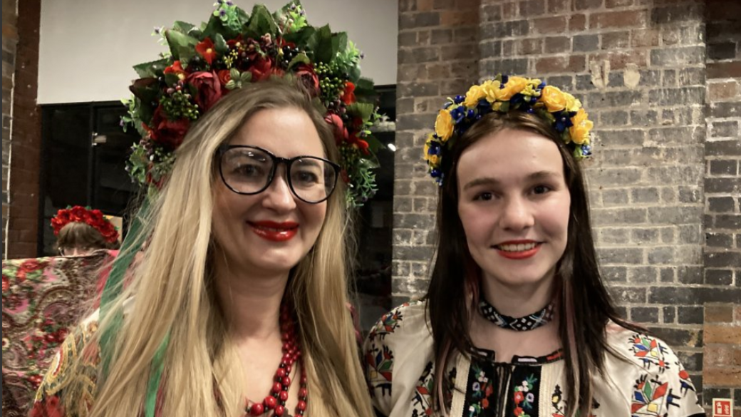 Two performers at the Ukrainian Christmas Service dressed in their national traditional costume during rehearsals. They have big floral head pieces, one woman is wearing black glasses and they have white or light coloured shirt-style tops with embroidered detailing. 