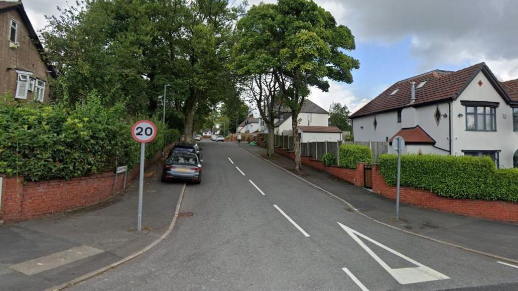 Woodgate Avenue with tall leafy trees and detached houses on either side. A road sign showing a 20mph speed limit stands on the corner.