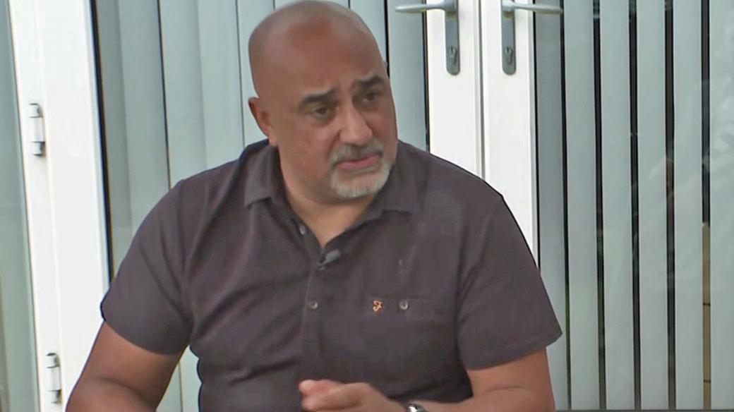Stephen Boston, in a dark, short-sleeved shirt sits in front of a patio window with blinds.