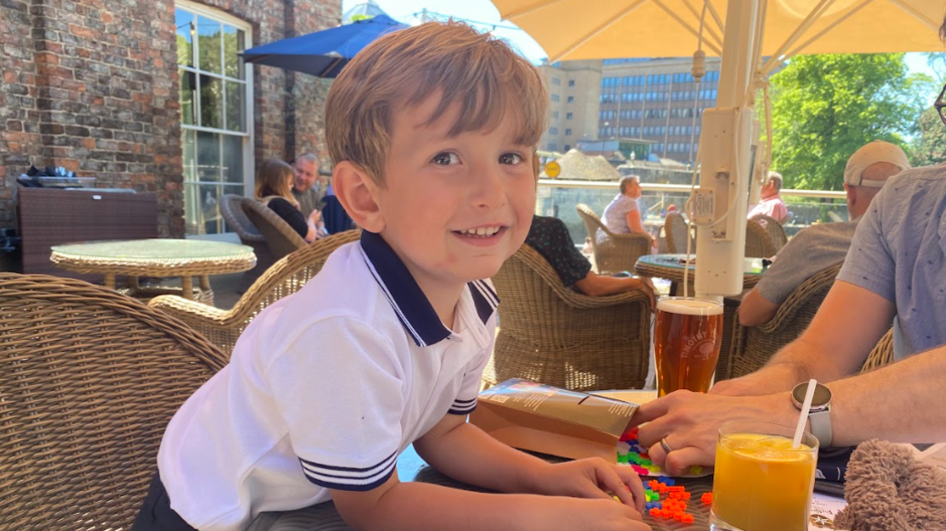Benedict is leaning across a table at an outdoor venue. He is smiling and has a white polo type sailors top on. He is playing with a game on the table where a pint of beer sits near a glass of organge juice.