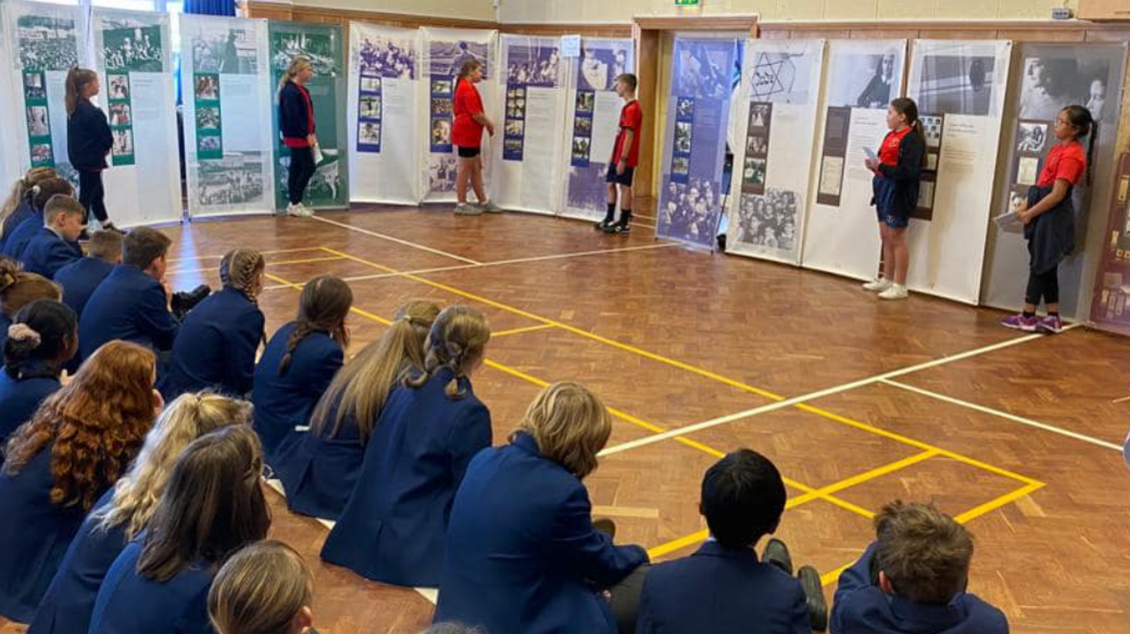 Children in blue blazers sitting on a brown wooden floor with their backs to the camera watching pupils share artwork and photographs from a history lesson.