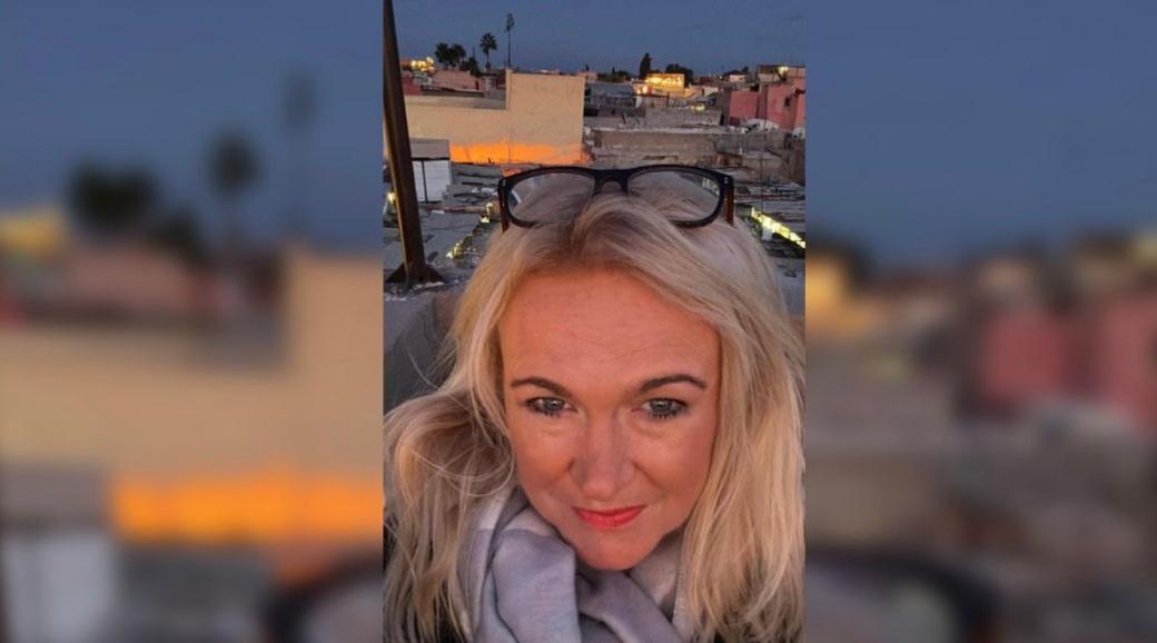 A selfie of Justine Tipling. She is in Marrakesh, Morocco. She has long blonde hair and has reading glasses with thick black frames on her head. The rooftops of buildings can be seen in the distance behind her.