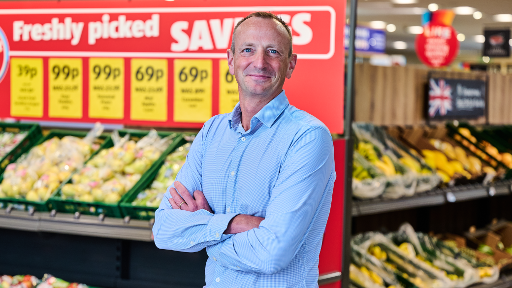 Giles Hurley, Aldi UK chief executive, poses with his arms folded in front of the fruit and veg aisle in an Aldi supermarket