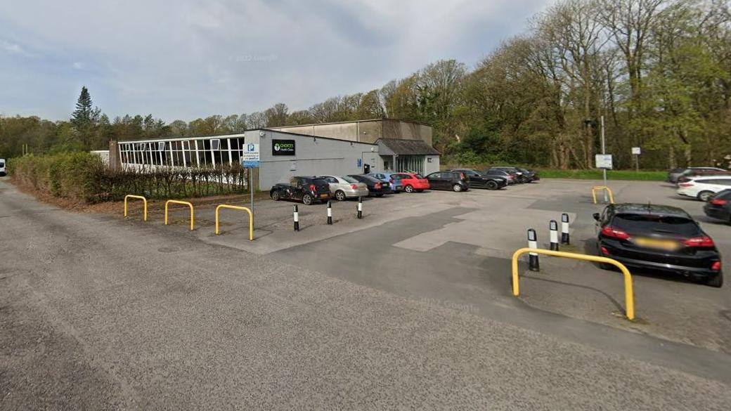 A Google Street screenshot of Troutbeck Bridge Swimming Pool. The building is a grey single-storey structure with a car park at the front. There is a line of large trees behind it and stretching away from the site.