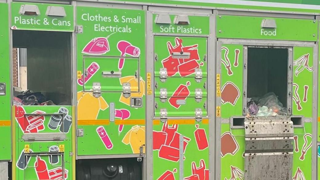 The side of a recycling truck with openings for plastics and cans, clothes and small electricals, soft plastics and food waste.
The truck is green with colourful drawings of everyday items in bright colours, pink, yellow and red, including hair dryers, clothes, bags, batteries, and pop bottles.
In the openings, you can see some bags of varied recycling.