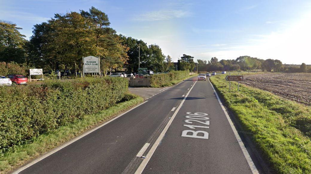 A Google Street View image of the B1206, Barton Road, near Elsham Golf Club. Cars are on the road travelling towards the camera. Grass verges line the right of the field. On the left of the road are bushes with an entrance to Elsham Gold Club and some cars in a car park.