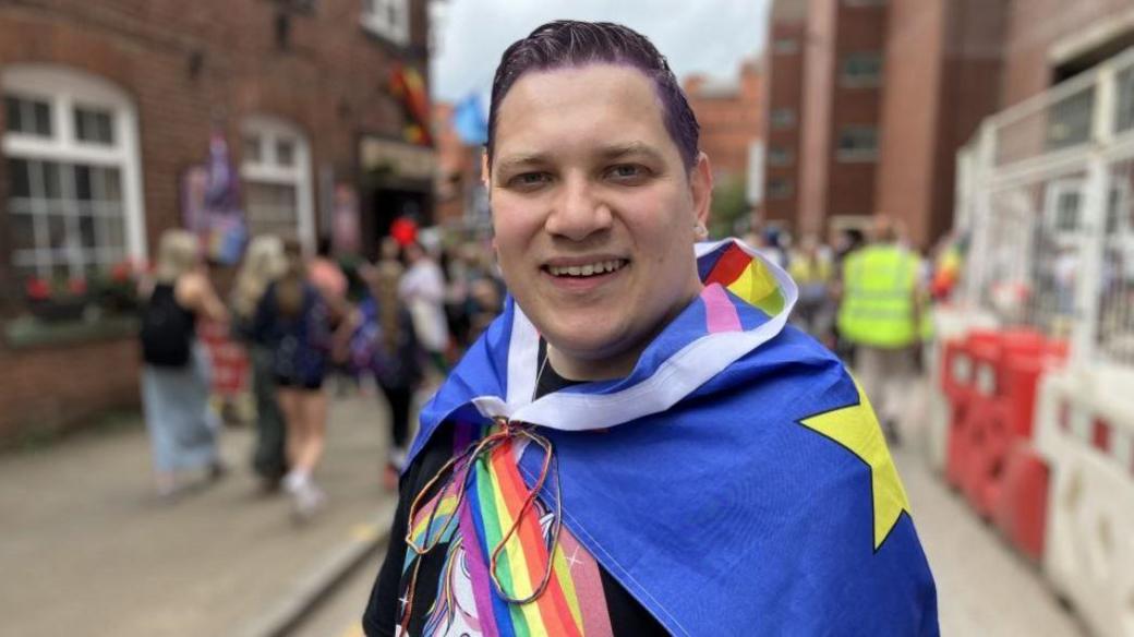 Organiser Councillor Louie Hamblett dressed in bright clothing and wearing a rainbow sash and a flag draped over him smiles into the camera with crowds behind him. He has purple hair.