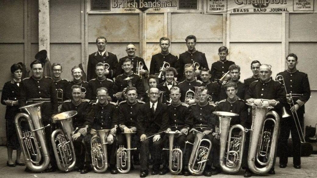 A black and white photograph of a historic brass band standing or sitting in rows holding their instruments.