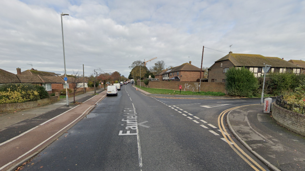 A tarmac road in a residential area. There is a road leading off to the right. Sky is cloudy. Brown brick houses and pathways line either side of the road