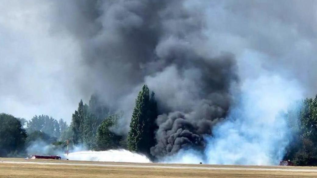 A huge billowing cloud of black, grey and white smoke coming from the edge of the airfield. It is so thick that it is hiding some of the trees - that are on the edge of the airfield. A fire engine can be seen spraying water towards the mass of smoke.