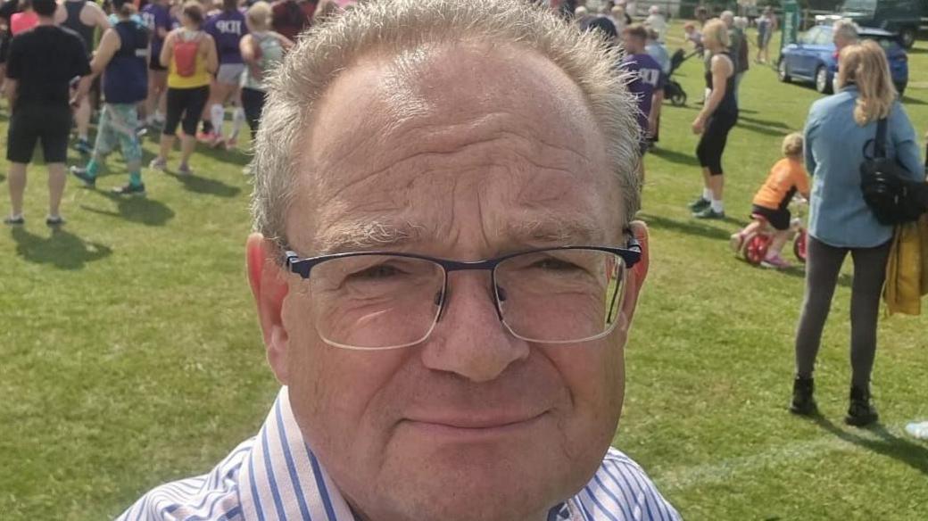 Jeremy Pert wears glasses and a striped shirt, he's in a field surrounded by people. It's a sunny day. He's out campaigning. 