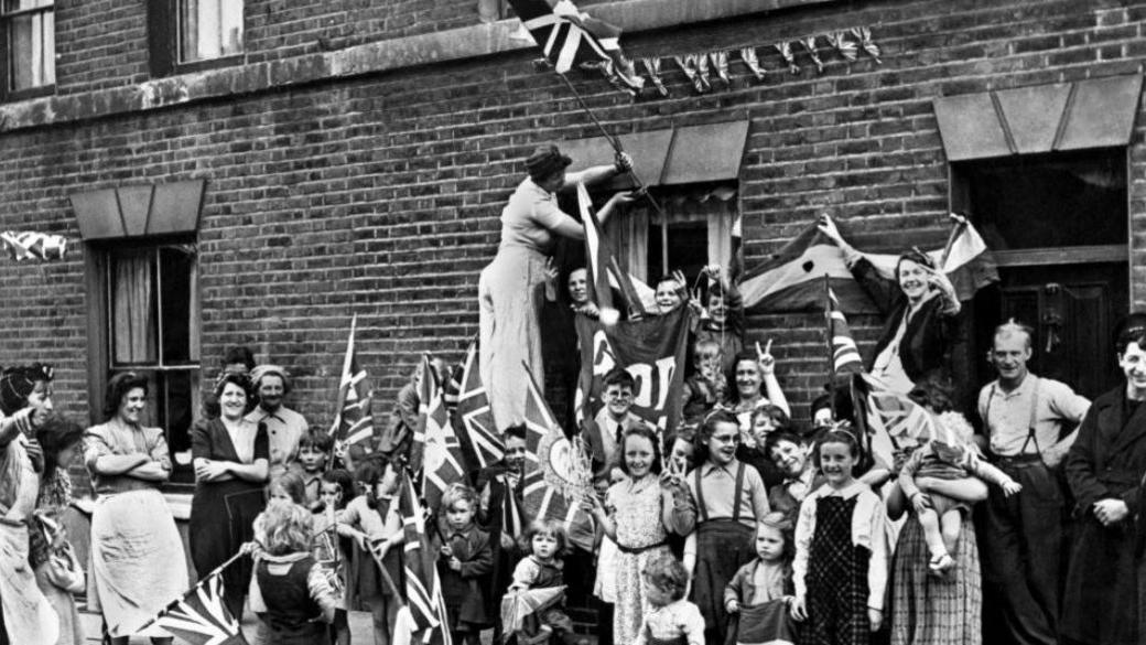 Men, women and children are celebrating VE Day in the street. In the background is a row of terraced houses adorned with flags.