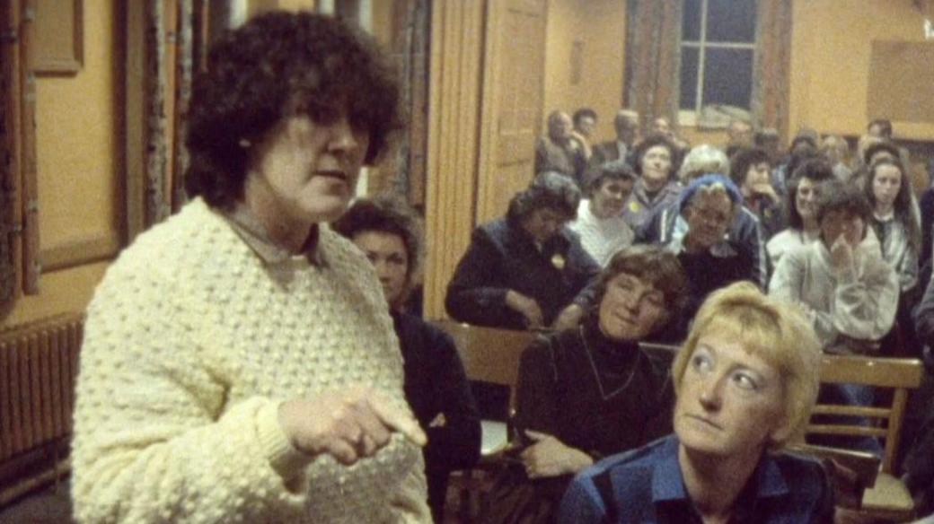 Margaret Matthews is standing to the left of frame wearing a cream wooly jumper. She has dark curly hair and she's pointing her finger. On her left is Kath Francis who has short blonde hair and is wearing a navy shirt. Behind them are a number of other women sitting down on wooden chairs in a hall which has mustard coloured walls.