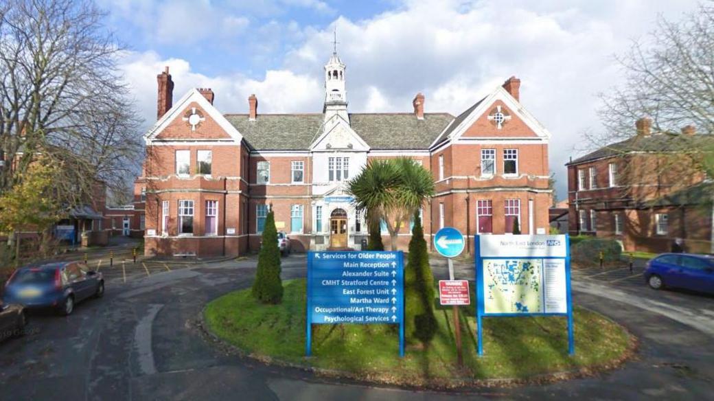 Goodmayes Hospital. Red brick building. Small grass roundabout in front with a blue sign and site map.