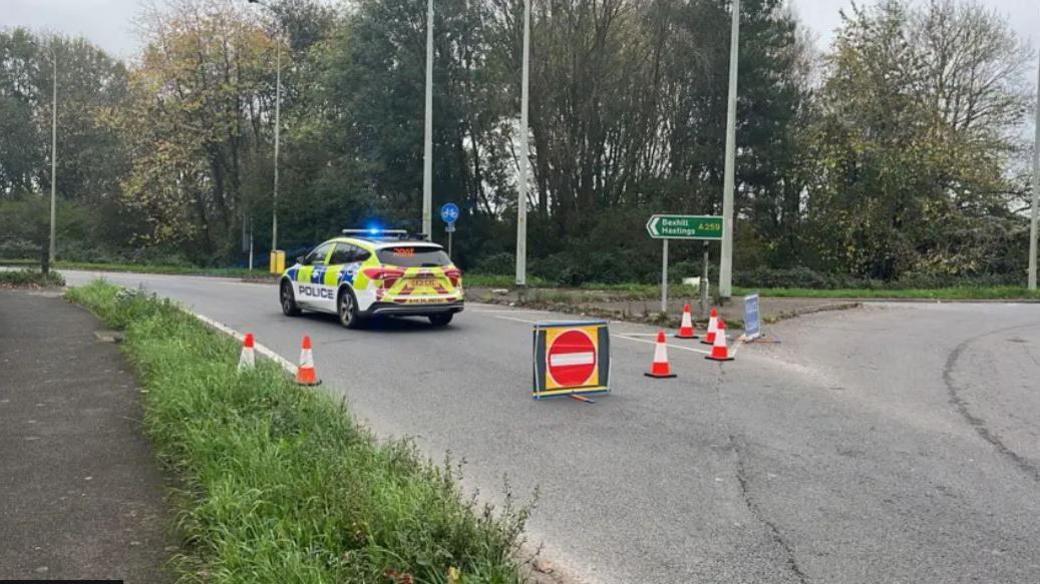 A police car at a road junction which has been closed off by cones and a stop sign. There is a sign in the background which reads A259 Bexhill Hastings 