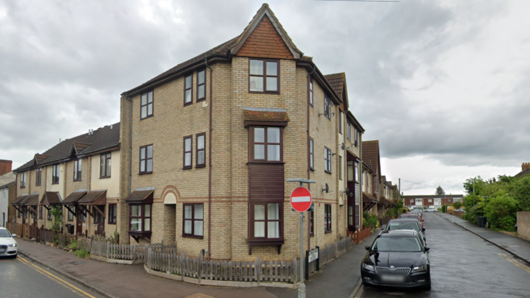 A block of brown-brick flats on a corner where two streets meet. Rows of houses lead to the block. Cars are parked in both streets.