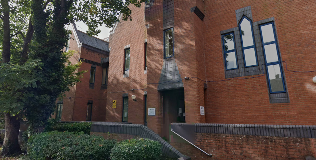 A red brick building with trees and a bushes outside and reflective windows.