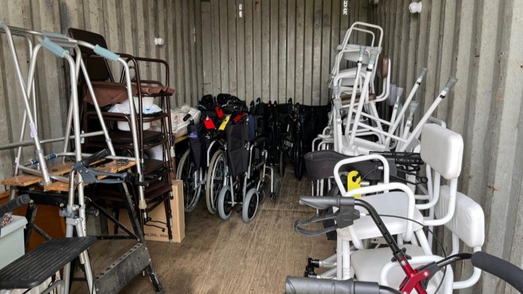 Mobility equipment is stored in a large metal container. There are various wheelchairs, walking aids and commodes.