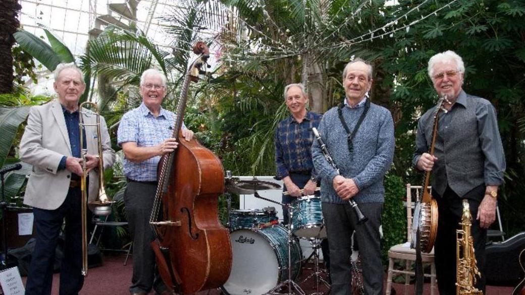 Jazz band The Savoy Jazzmen standing with their instruments smiling in an atrium.