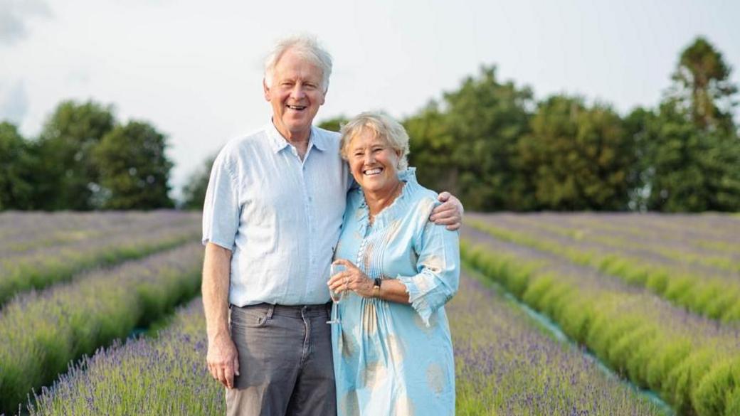Ian Jamie owns the farm with his wife Pippa and the couple are seen here celebrating their 40th wedding anniversary with a glass of bubbly in a lavender field. Both are wearing light blue and they are standing together looking happy.