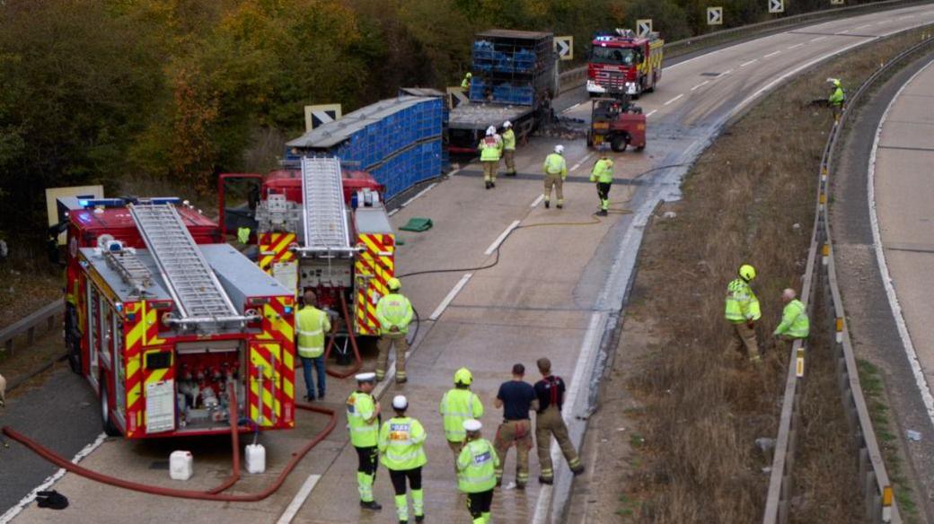 Fire engines and emergency workers on the A12. The picture is taken from above, from a bridge. A large blue container has been removed from a truck and is at the side of the road