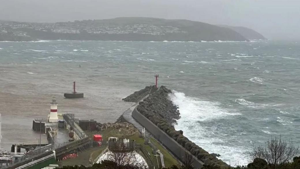 Grey skies over a choppy Douglas Bay, with waves crashing on the breakwater and Onchan Head in the distance.