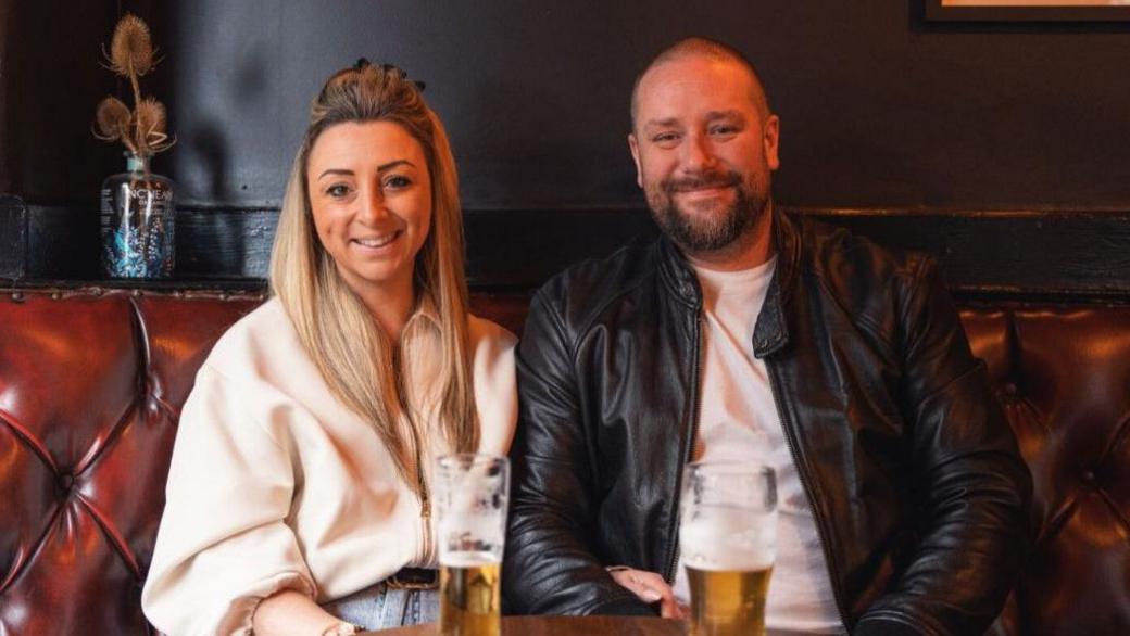 A woman with long blonde hair and a man with very short hair and a beard seated side by side in a pub setting on a brown sofa. They are both smiling and in front of them are two pint glasses of beer on a table. The background features dark walls.