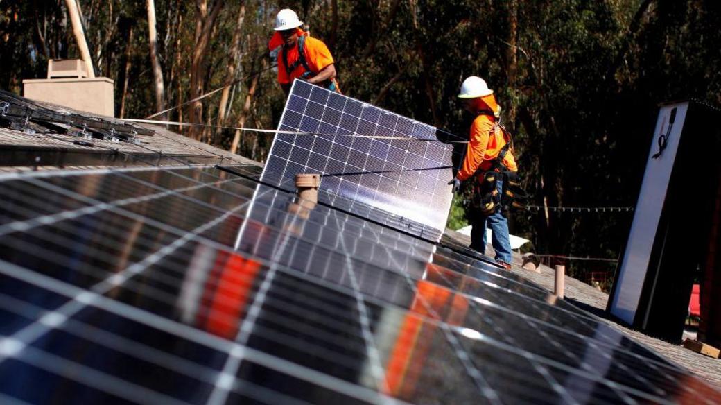 A solar panel is being installed by two employees on the roof of a property. They are wearing an orange high vis, a white hard hat and work trousers with boots.