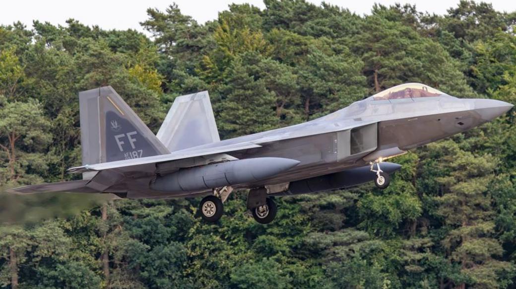 A grey US F-22A fighter jet coming into land. The aircraft is coming in low and green foliage can be seen behind it. The plane's undercarriage is visible with wheels out.