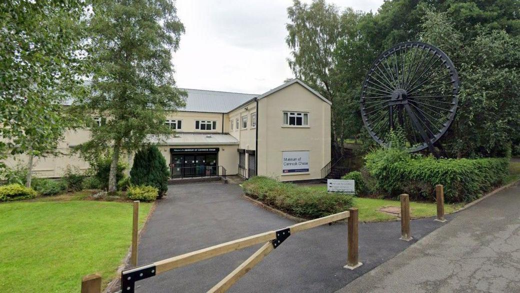 A cream-coloured two storey, l-shaped building set back from a single armed wooden gate and short tarmac driveway. To the left of the gate there is a preened lawn with trees and small bushes. To the right, a miner's wheel is elevated on a small mound. 