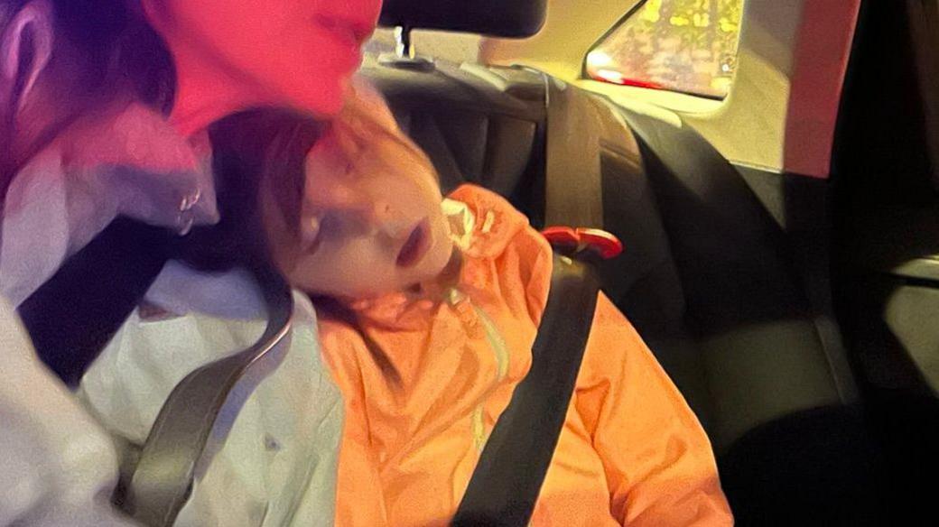 A young girl asleep in the back of a car. Her head is resting on the shoulder of a woman. She is wearing an orange coat and she has a seat belt on.