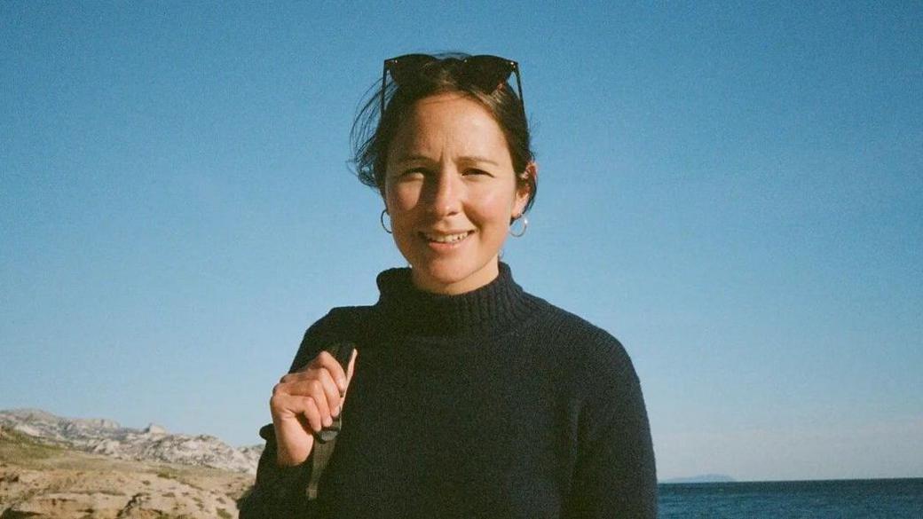 Daisy Moseley. She is pictured on the coast, with the sea behind her. She has dark hair and is wearing sunglasses on her head and a dark jumper. She is squinting slightly but looking at the camera and smiling. 